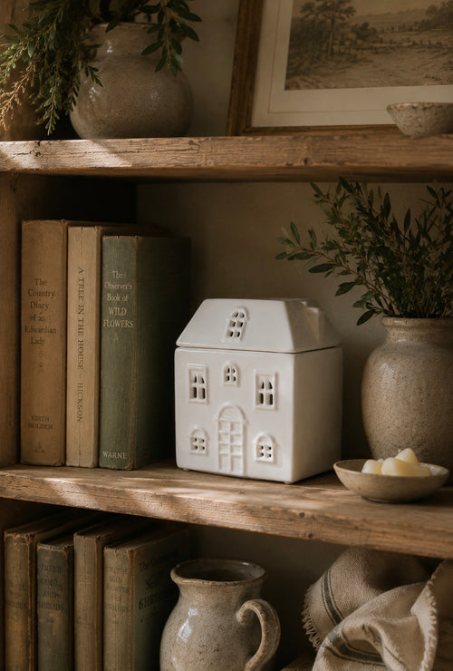 A white ceramic house-shaped container with window and door cutouts sits on a rustic wooden shelf beside books, a vase with greenery, and a small dish of candles.